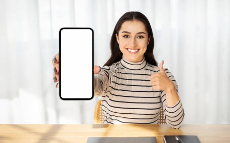 Photo for Millennial smiling arab lady show phone with blank screen, show thumb up gesture with hand, in room interior. App, blog recommendation, work or study website approve sign - Royalty Free Image