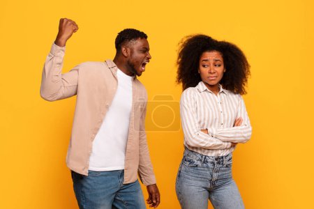 Téléchargez les photos : Homme noir criant agressivement avec le poing levé comme femme mécontente se tient avec les bras croisés, à la fois sur fond jaune soulignant la tension relationnelle - en image libre de droit