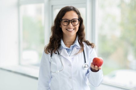 Téléchargez les photos : Belle jeune diétologue européenne portant des lunettes tenant la pomme à la main, recommandant des fruits frais pour une alimentation saine, la nutrition, posant à côté de la fenêtre à la clinique, espace de copie - en image libre de droit