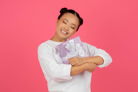 Joven dama asiática sonriente abrazando envuelto caja de regalo, retrato de alegre feliz coreano femenino sosteniendo presente, ojos cerrados en deleite, de pie aislado en el fondo del estudio rosa, espacio de copia