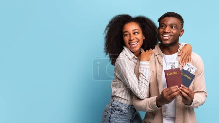 Foto de Joven pareja negra alegre sosteniendo pasaportes y tarjetas de embarque, anticipando ansiosamente su próximo viaje, mirando a un lado el espacio libre contra el calmante telón de fondo azul - Imagen libre de derechos