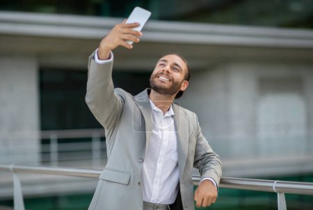 Photo for Joyful young businessman in suit taking a selfie outdoors, handsome smiling male entrepreneur capturing photo moment with his smartphone, posing at camera, displaying cheerful demeanor - Royalty Free Image