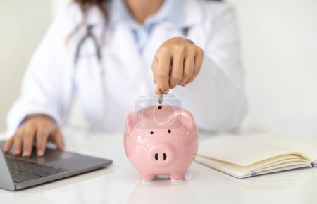 Cropped woman doctor general practitioner hand putting coin in piggy bank, managing hospital budget using laptop accounting applications, medical insurance finances, healthcare money savings concept