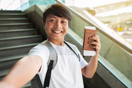 Foto de Alegre millennial asiático hombre bloguero de viajes tomando selfie en el aeropuerto o estación de tren, mostrando pasaporte y billetes de avión, sonriendo a la cámara. Viajar, turismo, bloguear - Imagen libre de derechos