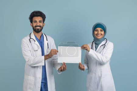 Téléchargez les photos : Médecins musulmans arabes confiants en blouse blanche tenant une pancarte blanche, jeunes travailleurs médicaux debout sur un fond de studio bleu, montrant l'espace de copie pour votre annonce, maquette - en image libre de droit
