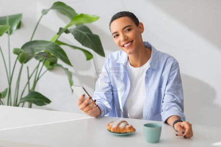 Photo for Relaxed young woman with a joyful smile using her smartphone at a white table, with a tasty croissant and a cup of coffee, indoor plants softly blurred in the background - Royalty Free Image