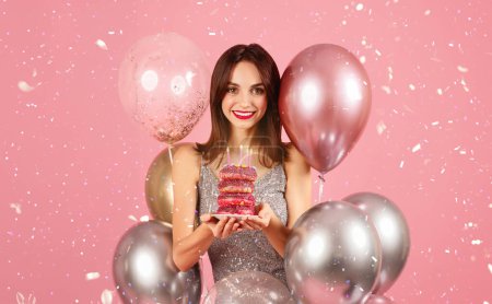 Photo for Joyous woman with a beaming smile celebrating with donuts topped with birthday candles, surrounded by a festive array of balloons amidst falling confetti on a pink background - Royalty Free Image
