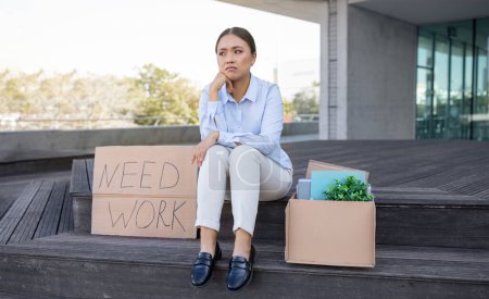 Téléchargez les photos : Chômage et licenciement. Femme d'affaires asiatique déprimée avec boîte en carton de biens et affiche Besoin de travail, assis à l'extérieur du bureau urbain, symbolisant la crise de carrière et la perte d'emploi - en image libre de droit