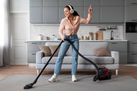 Photo for With headphones on, joyful woman dances while vacuuming her living room, turning mundane chore into an enjoyable and rhythmic experience - Royalty Free Image