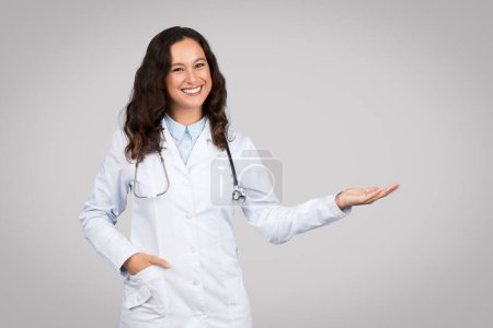 Confiada joven doctora en bata blanca y estetoscopio sonriendo, haciendo gesto de presentación con una mano sobre fondo gris, espacio libre