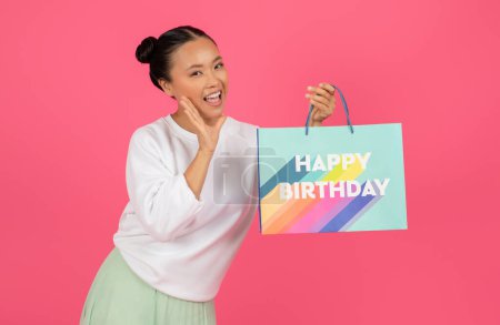 Cheerful young asian lady holding paper bag labeled Happy Birthday and touching face with excitement, happy korean woman ready to share joy at party, standing isolated on pink studio background