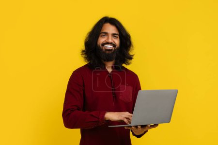 Téléchargez les photos : Éducation en ligne, apprentissage en ligne. Joyeux jeune étudiant indien barbu à distance utilisant un ordinateur portable, tapant sur le clavier de l'ordinateur, regardant la caméra et souriant, fond de studio jaune - en image libre de droit