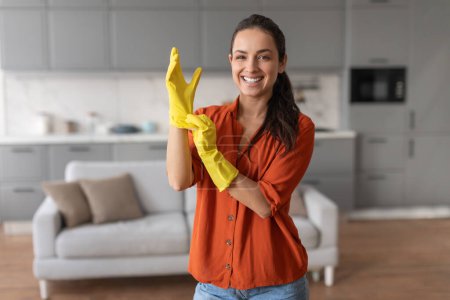 Téléchargez les photos : Femme joyeuse en chemise orange vibrante glisse sur des gants en caoutchouc jaune vif, se préparant à commencer ses tâches de nettoyage ménager avec le sourire - en image libre de droit