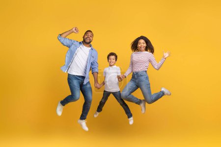 Téléchargez les photos : Portrait de joyeuse famille afro-américaine excitée de trois mains tenant tout en sautant dans l'air sur fond jaune en studio, heureux parents noirs et fils Preteen s'amusant ensemble, Copier l'espace - en image libre de droit