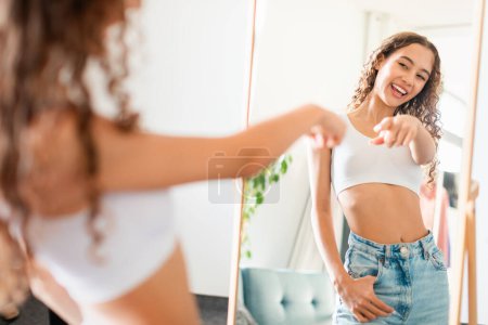 Photo for Young teen lady smiling at her reflection, pointing finger at mirror with confidence, celebrating body positivity on casual day at home. Selective focus, empty space - Royalty Free Image