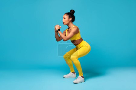 Photo for Workout. Fit African American Lady In Yellow Sportswear Doing Deep Squat Exercise During Work Out Practice Over Blue Studio Background, Wearing Earbuds And Smartwatch. Side View Shot, Copy Space - Royalty Free Image