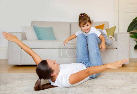 Photo for Mother and little daughter playing airplane game together at home, mother extending arms, pretending flying and lifting her delighted child on legs, having fun in living room interior - Royalty Free Image