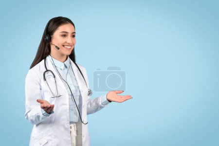 Friendly female doctor wearing a headset and gestures invitingly, prepared for an online teleconsultation or virtual assistance, blue background, free space
