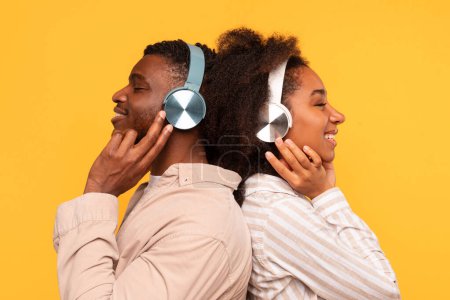 Photo for Back-to-back young black couple lost in music, each wearing and touching stylish headphones, profile view against vibrant yellow background - Royalty Free Image