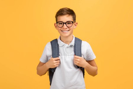 Téléchargez les photos : Joyeux écolier portant des lunettes et un sac à dos debout en toute confiance sur fond de studio jaune, heureux adolescent mâle prêt pour l'école, profiter de l'apprentissage et de l'éducation, copier l'espace - en image libre de droit
