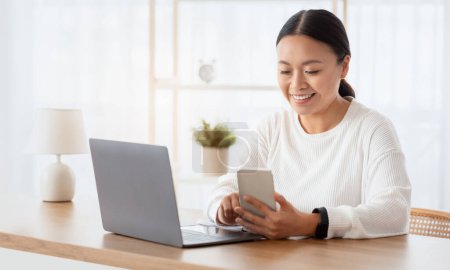 Téléchargez les photos : Femme asiatique joyeux entrepreneur travaillant de la maison, multitâche, assis au bureau, à l'aide d'un ordinateur portable et smartphone, répondre aux e-mails, vérifier l'application d'affaires, copier l'espace - en image libre de droit