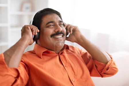 Foto de Primer plano del hombre indio de mediana edad feliz con bigote usando camisa naranja relajándose en el sofá en casa, usando auriculares inalámbricos, escuchando música favorita, mirando el espacio de copia - Imagen libre de derechos