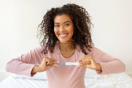 Photo for Happy beautiful black woman holding positive pregnancy test while sitting on bed at home, joyful young african american female expressing excitement about the new beginning, closeup shot, free space - Royalty Free Image