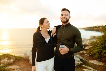 Foto de Hombre y mujer alegres en ropa deportiva, compartiendo momento de luz sosteniendo la botella de agua y abrazándose contra el fondo del atardecer en la playa - Imagen libre de derechos