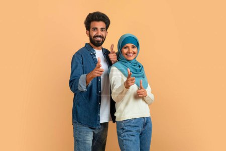 Gotcha. Fröhliches muslimisches Paar zeigt mit den Fingern in die Kamera, zeigt jemanden an, glücklicher arabischer Mann und Frau im Hijab, die zusammen Spaß haben, steht über beigem Studiohintergrund, Kopierraum