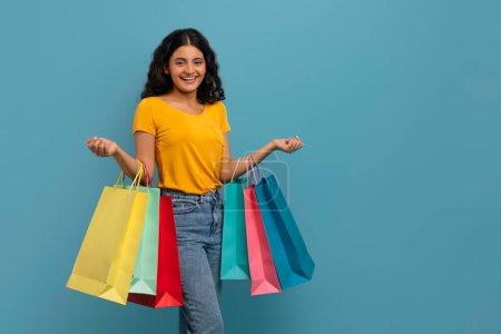 Excited happy attractive curly young indian woman student holding colorful shopping bags in both hands and smiling at camera, isolated on blue studio background, copy space. Consumerism, retail