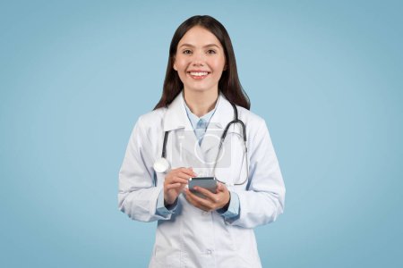 Cheerful female doctor engaged in using a smartphone, reflecting the integration of mobile technology in modern medical practice, on blue background