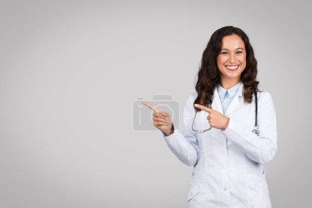 Téléchargez les photos : Médecin femme rayonnante en blouse blanche pointant joyeusement vers le côté à l'espace libre avec les deux mains, stéthoscope autour du cou sur un fond gris - en image libre de droit