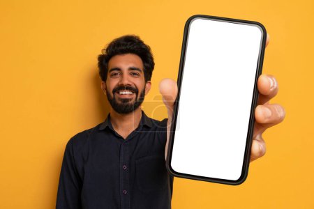 Téléchargez les photos : Homme indien joyeux montrant Big Phone avec écran blanc Mockup Copier de l'espace pour appareil photo, Publicité Grande offre en ligne tout en posant sur fond jaune Studio. Application mobile - en image libre de droit