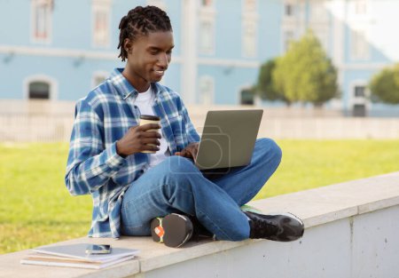 Foto de Feliz estudiante negro que estudia en línea usando el ordenador portátil, preparándose para los exámenes al aire libre, sentado en parapeto con taza de café de papel, websurf Internet mientras se aprende cerca del campus universitario - Imagen libre de derechos