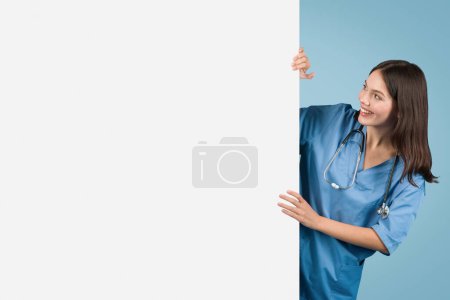 Téléchargez les photos : Infirmière joyeuse en tenue bleue jette un coup d'oeil ludique autour de grand panneau vierge, parfait pour l'information de santé ou la publicité sur un fond bleu, espace libre - en image libre de droit