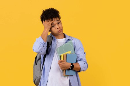 Photo for Contemplative young black male student with notebooks looks worried, hand on head, reflecting on problem against bright yellow, one-tone background - Royalty Free Image