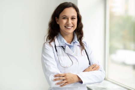 Téléchargez les photos : Portrait de joyeux jeune femme européenne brune médecin portant un uniforme médical blanc et stéthoscope posant à la clinique ou à l'hôpital à côté de la fenêtre, souriant à la caméra - en image libre de droit