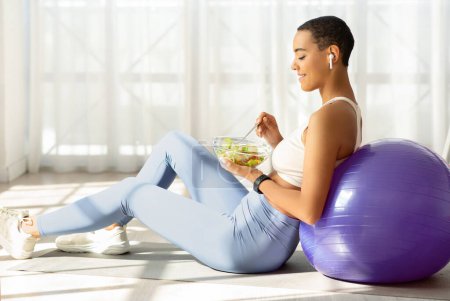Fit glücklich lateinamerikanische Frau genießen einen gesunden Salat, während sie auf einem Ball in einem sonnigen Raum sitzt, Ernährung und Fitness für einen ausgewogenen Lebensstil zu Hause kombiniert, genießen Sie das Mittagessen