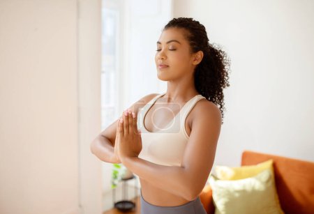 Photo for Sporty lady meditating with hands clasped standing in the calm of her living room, embodying mindfulness and balance in her morning yoga practice, posing with eyes closed - Royalty Free Image
