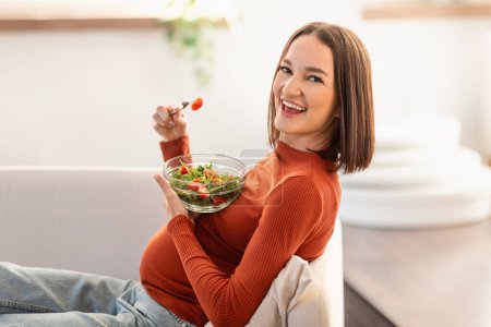Foto de Nutrición durante el embarazo. Feliz joven embarazada comiendo ensalada de verduras mientras está sentada en el sofá en casa, disfrutando de alimentos saludables con vitaminas orgánicas y relajándose en la sala de estar - Imagen libre de derechos