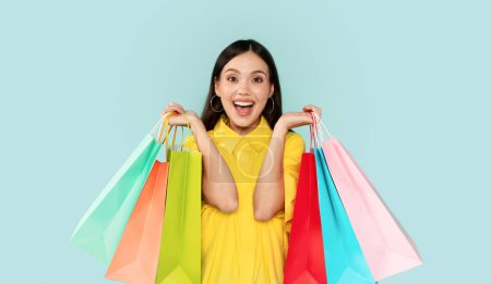 Emotional excited pretty brunette young woman wearing yellow shirt showing colorful shopping bags, buying clothes, cosmetics accessories during sale season, blue studio background