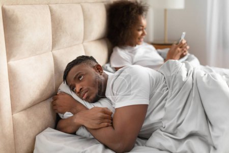 Infidelity, jealousy issue. Jealous black husband discontented about wifes online chats while she texting on cellphone, man suspecting cheating lying in bed in domestic bedroom. Selective focus