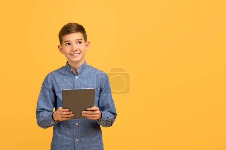 Téléchargez les photos : Jeune adolescent souriant tenant tablette numérique et regardant l'espace de copie, heureux adolescent masculin avec gadget moderne dans les mains debout sur fond jaune studio, penser à une offre agréable, Panorama - en image libre de droit