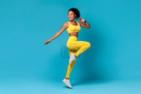 Photo for Young black woman in yellow sportswear jumping doing knee to elbow exercise on blue studio backdrop, displaying athletic motivation during workout. Healthy sport training - Royalty Free Image