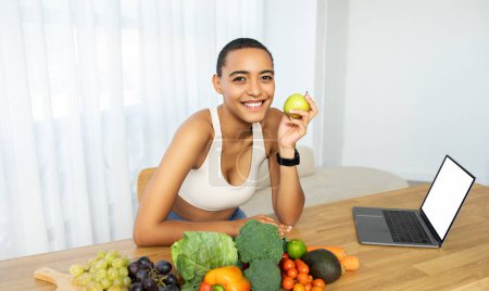Fröhliche lateinische Frau hält grünen Apfel mit einer Vielfalt an frischem Obst und Gemüse auf dem Tisch und diskutiert über gesunde Lebensweise am Laptop mit leerem Bildschirm zu Hause