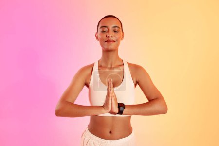 Téléchargez les photos : Une femme sereine avec une tête rasée pratique le yoga dans une pose paisible, les mains en anjali mudra, portant un soutien-gorge de sport et un pantalon lâche sur un fond rose et jaune dégradé - en image libre de droit