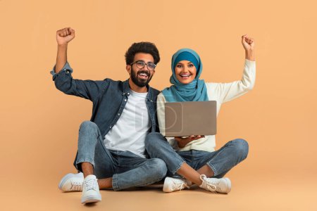Téléchargez les photos : Joyeux couple musulman arabe assis sur le sol avec ordinateur portable et célébrant le succès, heureux conjoints musulmans levant les bras dans la victoire, acclamant les bonnes nouvelles, posant sur fond de studio beige, espace libre - en image libre de droit