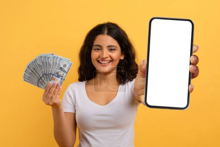 Photo for Happy beautiful young indian woman gambling on Internet, using smartphone with white empty screen, showing bunch of dollars cash, yellow studio background, mockup. Online bet concept - Royalty Free Image