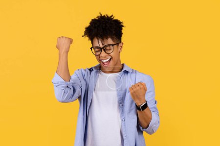 Téléchargez les photos : Jeune étudiant noir extatique avec des lunettes serre les poings dans la victoire, les yeux fermés dans l'exaltation, sur fond jaune vif, symbolisant triomphe - en image libre de droit
