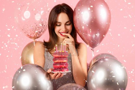 Téléchargez les photos : Charmante femme caucasienne millénaire au sourire discret, contemplant ludique une bouchée d'un gâteau au beignet rose empilé de bougies allumées, entouré de ballons et de confettis sur fond rose - en image libre de droit